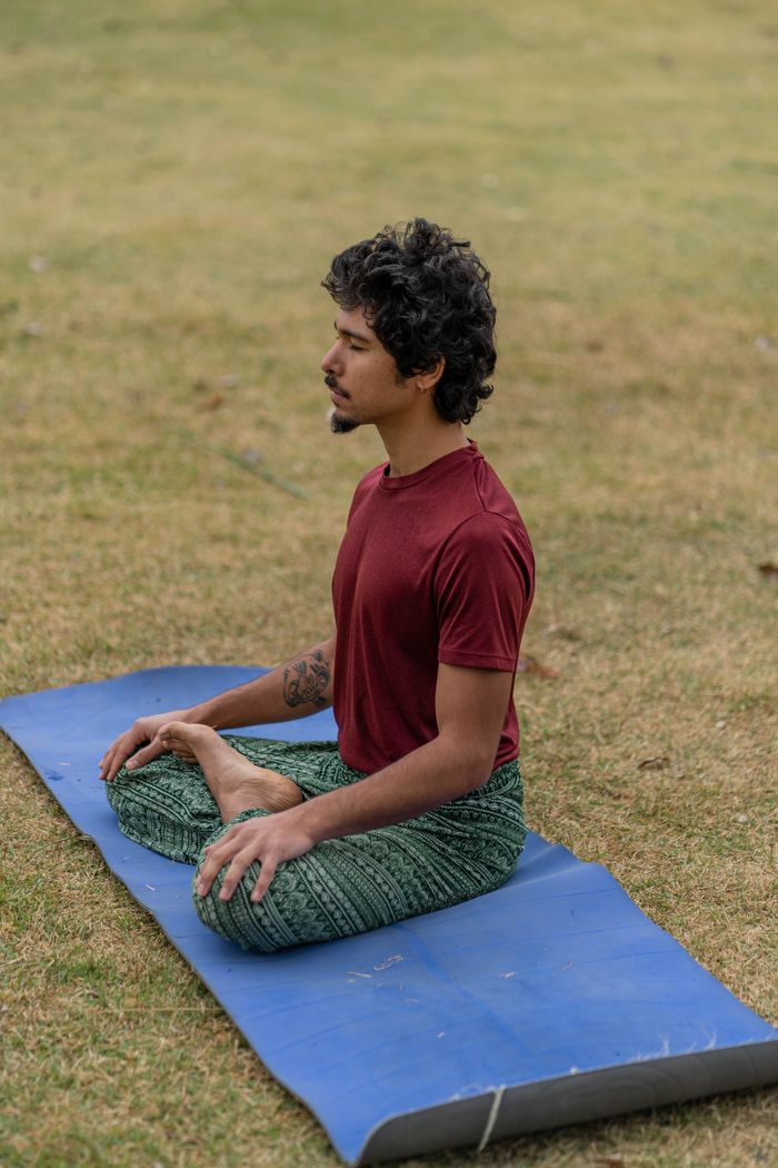 A man meditating in a relaxed posture on a blue yoga mat in a grassy field.